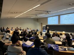 Image showing a man speaking to a group of students in a large odeon-style classroom. Text on the screens behind him reads "Welcome to the Master of Supply Chain Management Program"